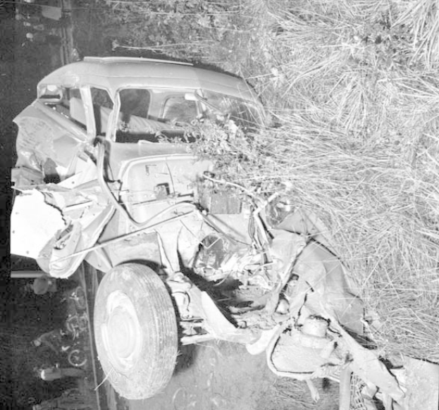 Schwarz-weiß-Foto eines schwer beschädigten Autos am Straßenrand mit eingedellter Front, zersplitterten Fenstern und verstreuter Trümmer im Hintergrund.
