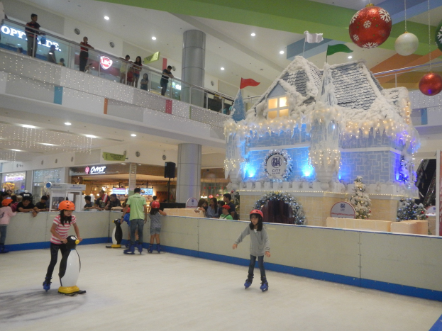 Menschen, die auf einer Eisbahn in einem Einkaufszentrum Schlittschuh laufen, umgeben von festlichen Dekorationen, darunter ein häuserähnlicher Aufbau mit Lichtern und eine Menge, die von verschiedenen Ebenen aus zusieht.
