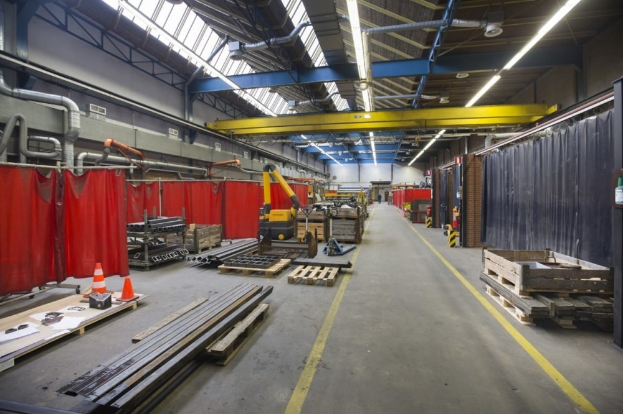 Ein großer Lagerraum mit Holzpaletten, Metallobjekten, Verkehrskegeln und Maschinen, mit roten Vorhängen links, einer Vorhangwand rechts und Deckenlampen und Rohren.