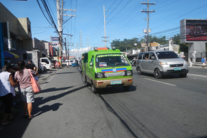 Ein grüner Lieferwagen fährt auf einer Stadtstraße mit hohen Gebäuden auf beiden Seiten, Passanten auf dem Gehweg, Strommasten mit Drähten, Bäumen im Hintergrund und einem klaren blauen Himmel.