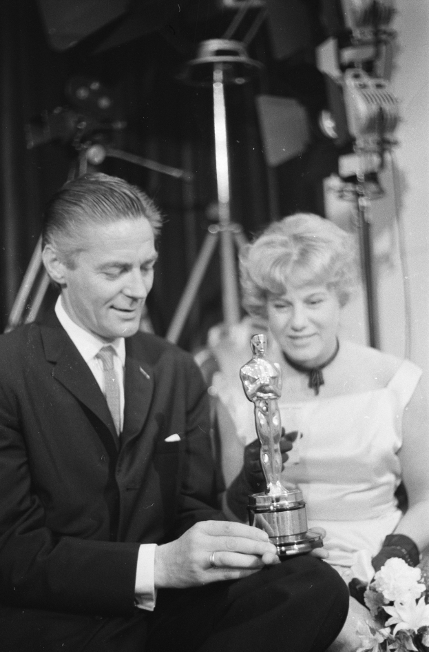 Ein Mann in einem schwarzen Blazer, weißem Hemd und Krawatte hölt eine Oscar-Statue, waß er neben einer Frau in Abendgarderobe sitzt, die einen Blumenstrauß hölt, mit weicher Beleuchtung im Hintergrund.