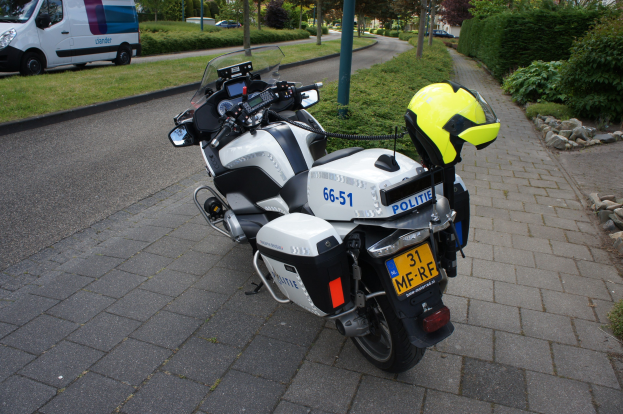 Eine Polizei-Motorrad auf dem Seitenstreifen geparkt mit einem Helm darauf, umgeben von Bäumen, Pfählen, Gebäuden, Pflanzen, Gras, Steinen und anderen Fahrzeugen.