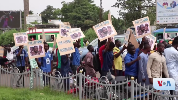 Eine Gruppe von Menschen mit Schildern steht vor einem Zaun, umgeben von Fahrzeugen, Bäumen, Pfählen, Plakaten und einer Leinwand, mit Himmel und Gras im Hintergrund, wahrscheinlich protestieren sie gegen eine Regierungsentscheidung, die Video-Konferenzen verbietet.
