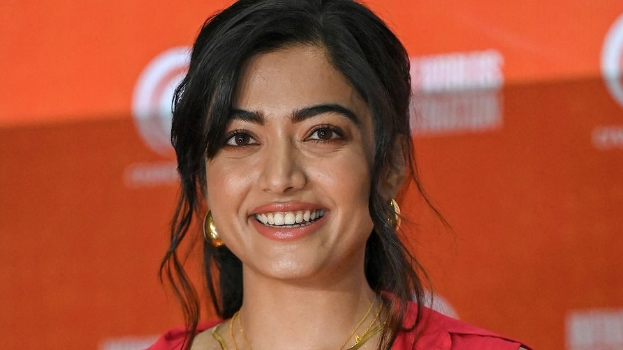 Eine Frau in einem roten Kleid mit einem eleganten Dutt und dezentem Make-up lächelt in die Kamera, trägt schwarze High Heels und hat einen hellgelben Banner mit der Aufschrift "Sonam Kapoors beste Schönheits Looks vom Cannes Film Festival" im Hintergrund.