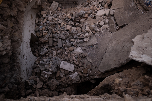 Zusammengebrochenes Gebäude mit einem großen Loch im Boden, umgeben von Trömmern und einer sichtbaren Rohrleitung auf der linken Seite, die Erdbebenschäden anzeigt.