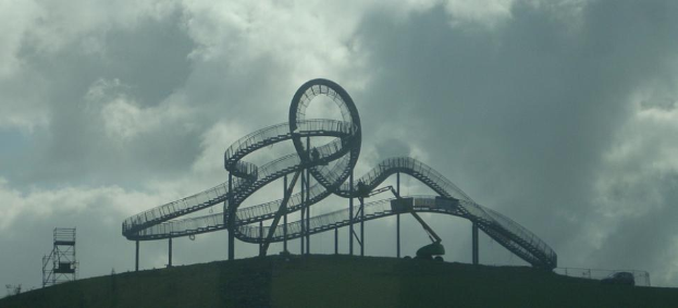 Eine Achterbahn auf dem Höhepunkt eines Hügels mit einem bewölkten Himmel im Hintergrund, umgeben von Stützpfählen und Fahrgeschäftsinfrastruktur.