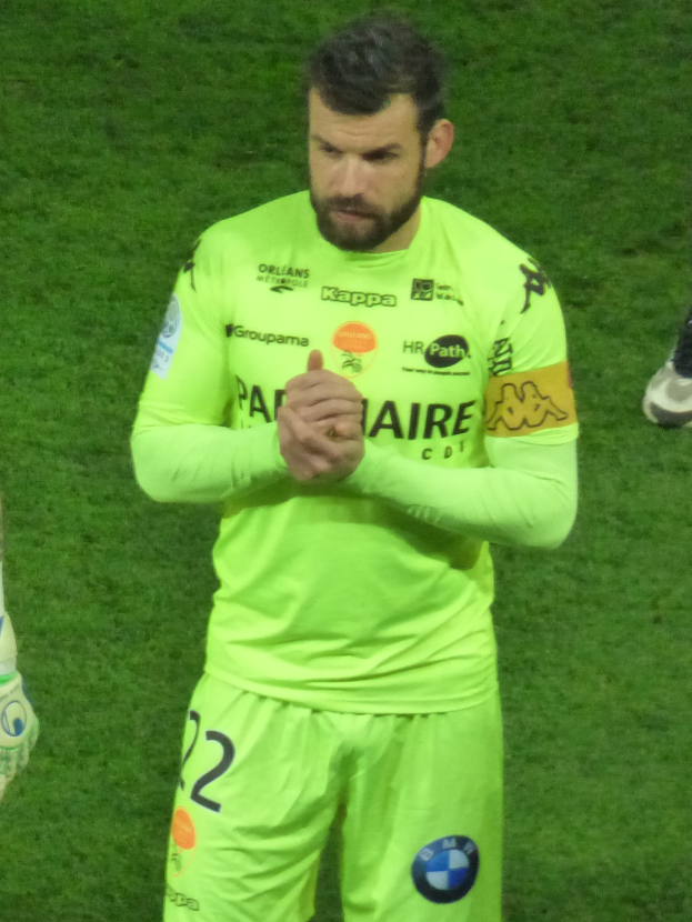 Ein Mann in einem gelben Torwarttrikot, der ein grünes T-Shirt und Shorts trägt, steht auf einem Fußballfeld und klatscht in die Hände, wobei auf der linken Seite des Bildes das Bein einer anderen Person zu sehen ist.