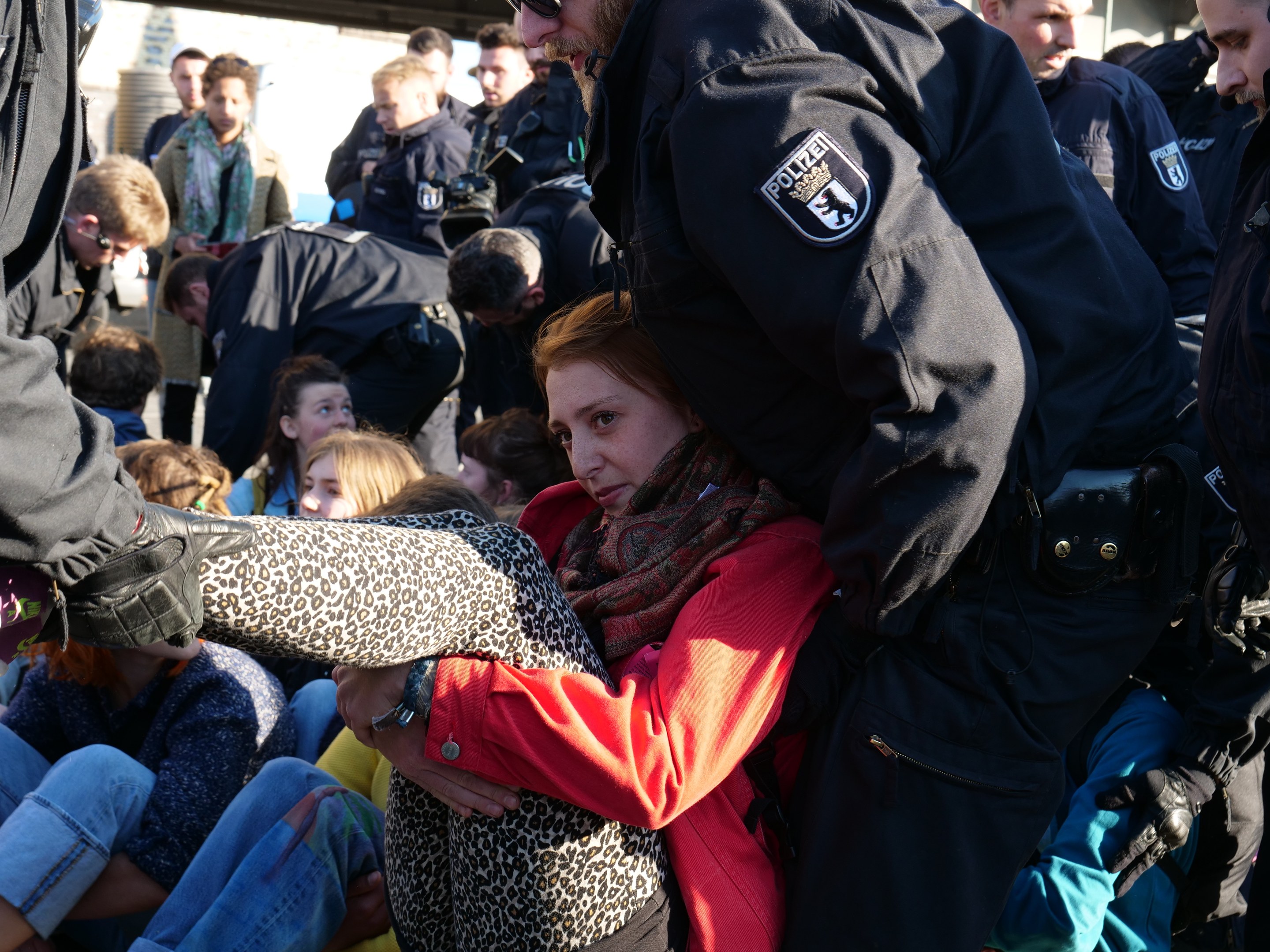Eine Frau in dunkler Kleidung wird von einem Polizeibeamten in Uniform begleitet, hinter ihnen sitzen und stehen Menschen, im Hintergrund ein Schuppen und ein klarer blauer Himmel.