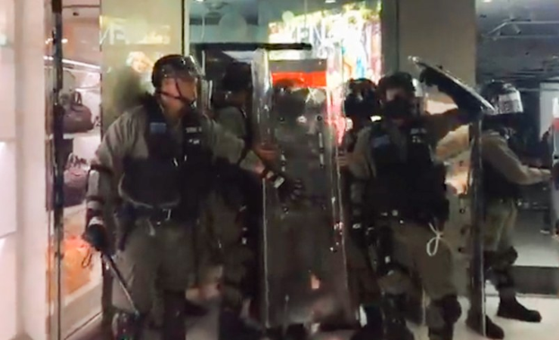 Gruppe von Polizisten in Uniform vor einem Geschäft stehend, mit Helmen und Gegenständen bewaffnet, vor Lichtern und anderen Gegenständen im Hintergrund und einer Wand auf der linken Seite.