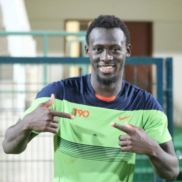 Ein Mann in einem grünen und blauen Hemd, der fröhlich in die Kamera zeigt und im Hintergrund eine undeutliche Reling, Wand und Tür zu sehen sind.