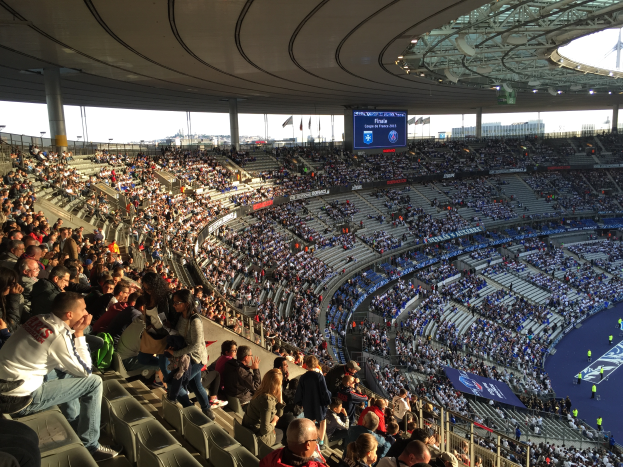 Eine große Menschenmenge, die in einem Stadion ein Fußballspiel schaut, mit einer Bühne auf der rechten Seite, Fahnen, Stangen und einem Bildschirm im Hintergrund, unter einem sichtbaren Himmel, in der Allianz Arena in München, Deutschland.