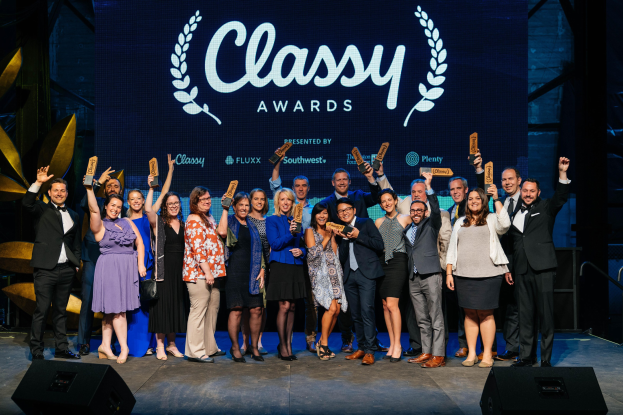 Group of people on a stage holding awards, with speakers below and a screen displaying 'Classy Awards 2019' in the background.