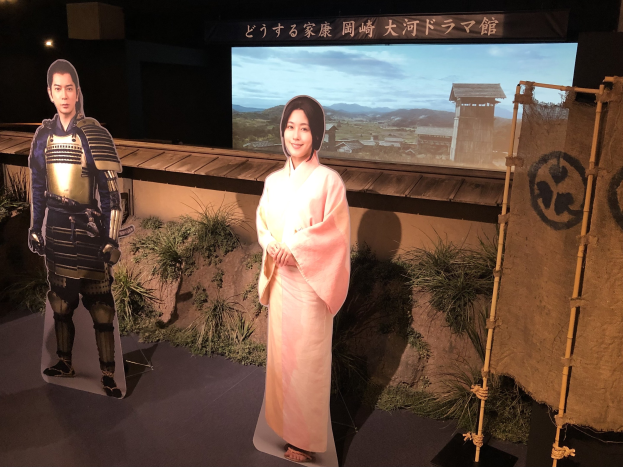 Zwei Personen in traditioneller japanischer Kleidung - eine Frau in einem pinken Kimono und ein Mann in Samurai-Rüstung - lächeln gemeinsam in einer Inszenierung mit Pflanzen, einem Schirm und Beleuchtung, die auf eine Filmszene hinweist.