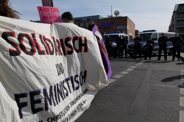 Gruppe von Menschen marschiert die Straße entlang und hölt eine 'Solidarität und Feminismus'-Fahne, mit geparkten Fahrzeugen und Gebäuden im Hintergrund bei klarem blauem Himmel.