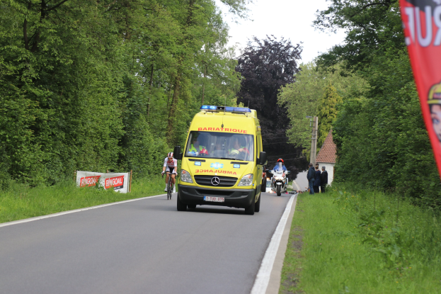 Ambulanz fährt auf Straße mit Radfahrern daneben, Gras und Bäume auf beiden Seiten, Häuser, Pfähle und einen klaren blauen Himmel im Hintergrund.