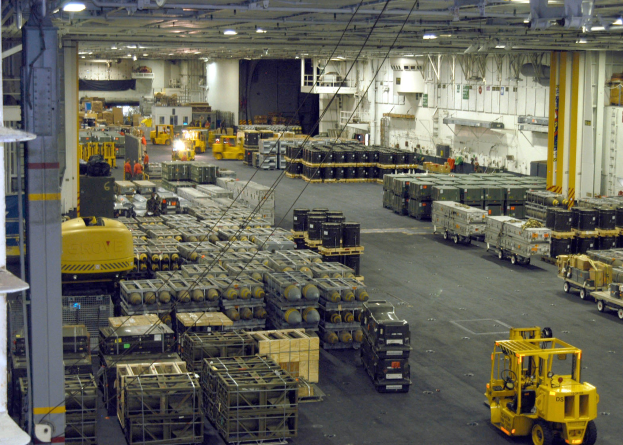 Lagerhalle mit Lkw, Containern, Menschen, Deckenbeleuchtung, Metallstangen und einem sichtbaren Rad.