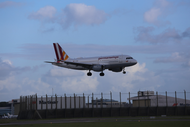 Germanwings Airbus A320-200 beim Starten am Frankfurter Airport mit Gras darunter, einem Zaun im Vordergrund, Gebäuden und Bäumen im Hintergrund und einer bewölkten Himmel über ihnen.