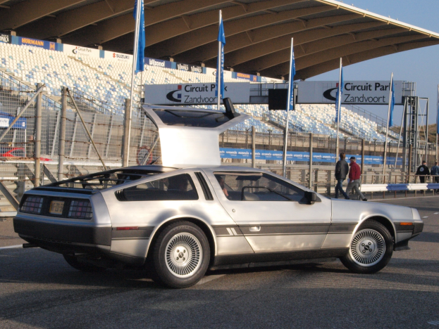 Ein DeLorean DMC-12 Auto auf einer Straße umgeben von Menschen, Fahnen, Schildern, Geländern, Stühlen und einem Schuppen, mit Bäumen und einem klaren blauen Himmel im Hintergrund.