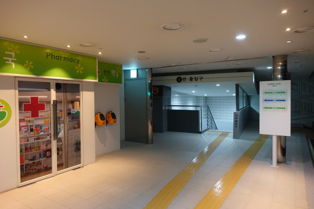 Apothekeneingang mit einem "Apotheke"-Schild links, einer Tafel mit Text rechts, Hintergrundobjekten und beleuchteter Dachbeleuchtung.