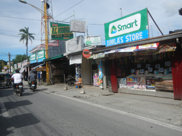 Eine belebte Straße mit Motorrädern, Autos, Gebäuden, Schildern, Strommasten, Drähten, Straßenlaternen, Bäumen und einem bewölkten Himmel.