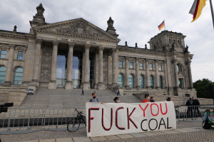 Menschen mit einem "Fuck You Coal"-Schild vor dem Reichstag in Berlin, mit Bäumen, einer Flagge und einem bewölkten Himmel im Hintergrund.