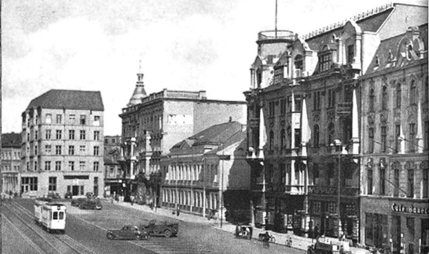 Ein Schwarz-Weiß-Foto einer Straßenkreuzung in Stuttgart, Deutschland, circa 1900, mit Fahrzeugen, Gebäuden mit Fenstern, Laternenpfählen und einem bewölkten Himmel.