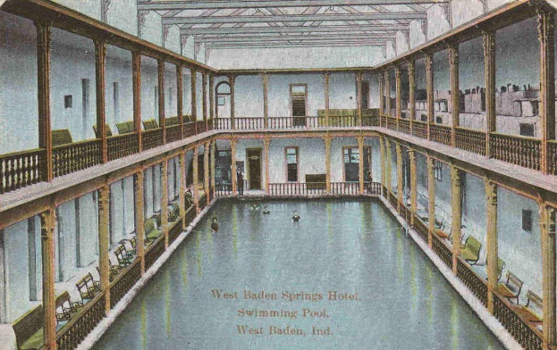Schwarz-weiß-Postkarte des großen Innen-Schwimmbads des West Baden Springs Hotels mit Menschen, umgeben von Säulen, Geländern, Stühlen und architektonischen Details, mit Text unten.