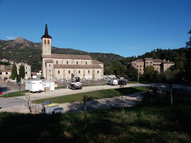 Kleiner Ort mit einer zentralen Kirche, umgeben von Gebäuden, Bäumen, Fahrzeugen und Schildern, vor einem Hügel- und einem klaren blauen Himmel.