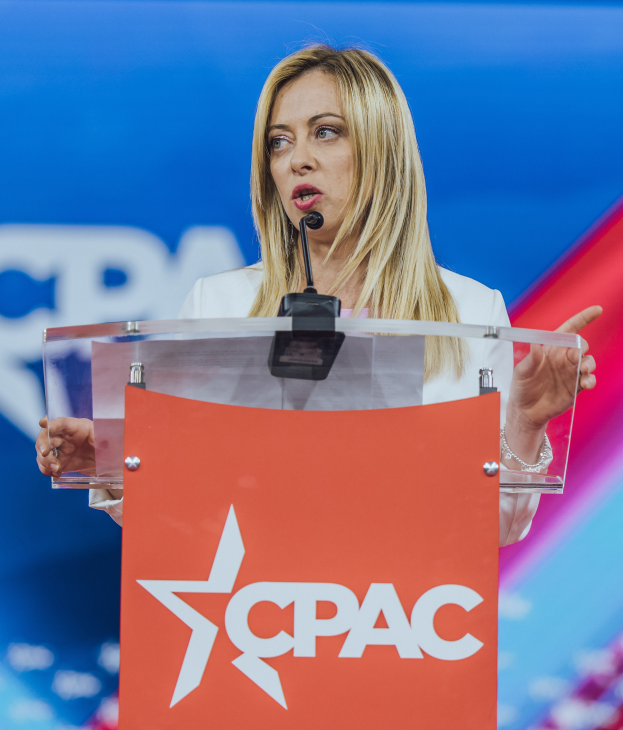 Eine Frau in einem weißen Kleid steht an einem Podium mit einem Mikrofon, die zu sein scheint eine politische Wissenschaftlerin, mit einem unscharfen Hintergrund.