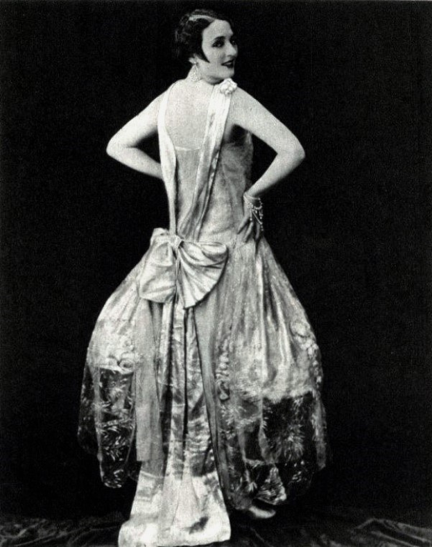 Eine Frau in einem Kleid im Stil der 1920er Jahre mit hohem Kragen, langen Ärmeln und einem weiten Rock steht selbstbewusst, ihre Haare sind elegant hochgesteckt und das Make-up natürlich.