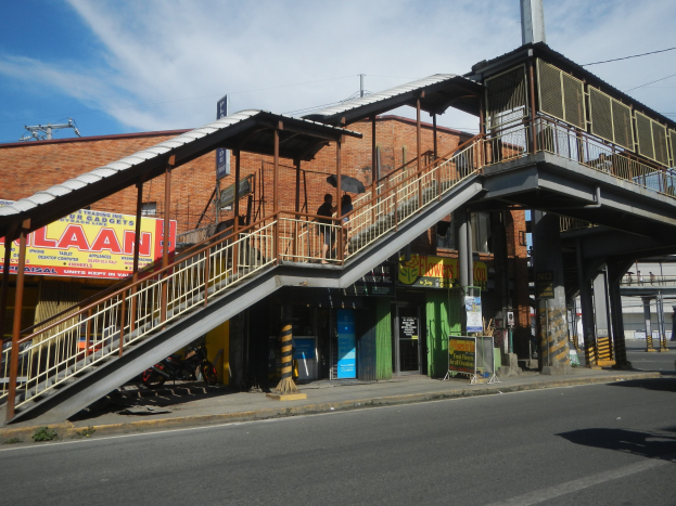 Ein Bahnhof mit einem Zug auf den Schienen, eine Brücke mit Geländern und Treppen, Gebäude mit Namensschildern, parkende Motorräder an der Straße, Strommasten mit Oberleitungen und ein bewölkter Himmel im Hintergrund.