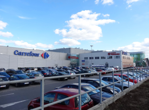 Großes Autohaus mit parkenden Fahrzeugen, umgeben von Gebäuden, Straßeninfrastruktur und Fahrzeugen auf der Straße unter einem bewölkten Himmel.