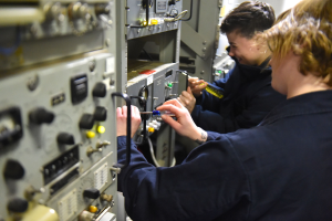 Zwei Männer in einer Fabrik, die an einer Maschine arbeiten und jeweils Werkzeuge halten, umgeben von elektronischen Geräten.