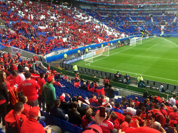 Eine große Menschenmenge in einem Stadion schaut einem Fußballspiel zu, mit Menschen auf Stühlen und Stehplätzen, Fahnen, einem Torpfosten und Deckenleuchten.
