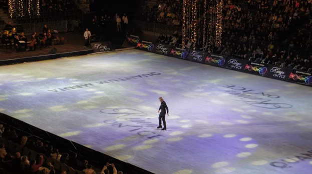 Ein Mann steht auf einer Eisbahn bei den Olympischen Winterspielen 2018 und performt vor einem sitzenden Publikum. Links von ihm stehen ein paar Menschen auf der Bühne und dekorative Lichter hängen über ihnen.