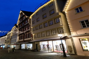 Headsteinstraße in Rothenburg ob der Tauber, Deutschland, geschmückt mit festlichen Weihnachtslichtern, mit Menschen, die spazieren gehen und ein geparktes Fahrrad, flankiert von Gebäuden mit Fenstern unter einem sichtbaren Himmel.