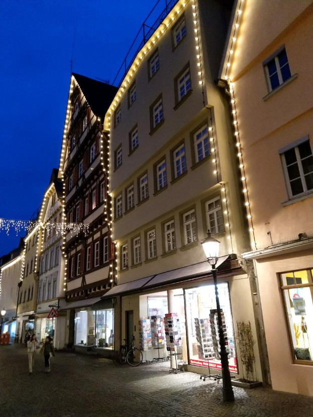 Headsteinstraße in Rothenburg ob der Tauber, Deutschland, geschmückt mit festlichen Weihnachtslichtern, mit Menschen, die spazieren gehen und ein geparktes Fahrrad, flankiert von Gebäuden mit Fenstern unter einem sichtbaren Himmel.