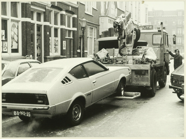 Ein Schwarz-Weiß-Foto eines Autos, das von einem Abschleppwagen auf einer Straße mit anderen Fahrzeugen, einer Person in der Nähe und Gebäuden mit Fenstern im Hintergrund geschleppt wird.