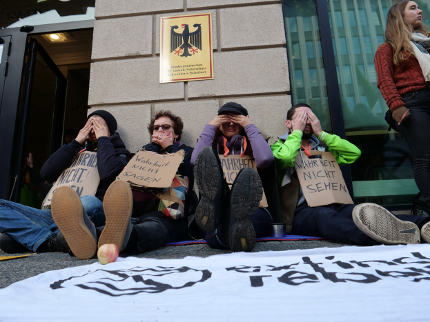 Gruppe von Menschen, die vor einem Gebäude auf dem Boden sitzen, Schilder halten, mit einem Banner auf der Straße und einer Frau rechts daneben; Hintergrund enthält eine Wand mit einer Tafel und ein Gebäude mit Glasfenstern und einer Tür.