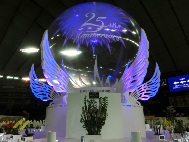 Ein großer Kristallball mit Flügeln oben drauf, umgeben von Blumenschmuck, Namensschildern und anderen Gegenständen auf dem Boden, mit Schildern, Lichtern und einem Bildschirm im Hintergrund bei einer 25-jährigen Jubiläumsfeier.