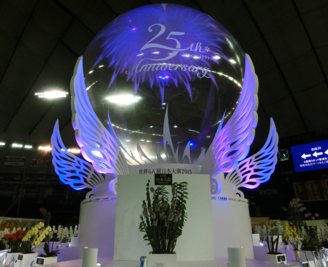 Ein großer Kristallball mit Flügeln oben drauf, umgeben von Blumenschmuck, Namensschildern und anderen Gegenständen auf dem Boden, mit Schildern, Lichtern und einem Bildschirm im Hintergrund bei einer 25-jährigen Jubiläumsfeier.