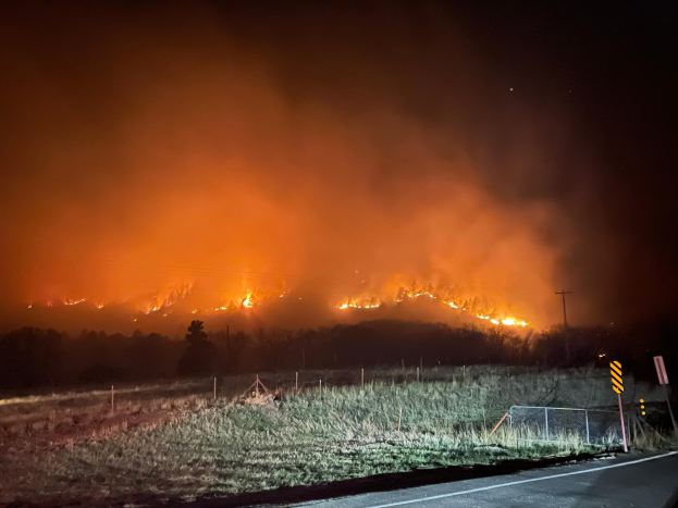Großer Brand in einem Feld in der Nacht mit Gras, einem Zaun, Pfosten und Bäumen im Vordergrund unter einem dunklen Himmel.