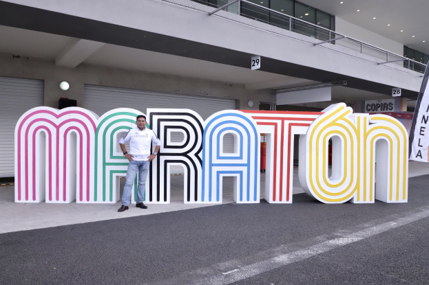 Ein Mann steht am Start eines Marathons vor einem großen "Marathon"-Schild, mit einem Banner rechts daneben und einem Gebäude mit Lampen, Geländern und Fensterläden im Hintergrund.