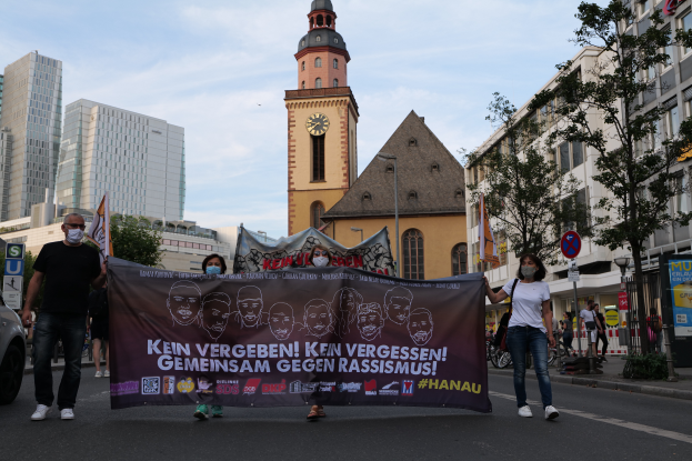 Eine Gruppe maskierter Menschen marschiert auf einer Straße und hält ein Banner hoch, mit einem geparkten Auto auf der linken Seite, Gebäuden, Bäumen, Schildern, Polen und einem Uhrenturm im Hintergrund unter einem klaren blauen Himmel.