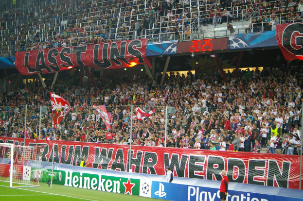 Fußballspiel in einem Stadion mit einer großen Zuschauermenge, saftigem Grün, einem Tor auf der linken Seite, Bodenbanner, Menschen mit Fahnen und Hintergrundgeländer, Tafeln, Lichter und andere Gegenstände.