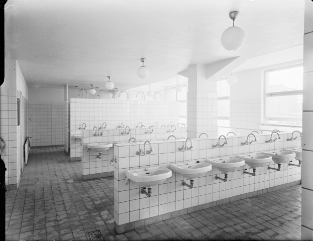 Ein Schwarz-Weiß-Foto einer öffentlichen Toilette mit mehreren Waschbecken, Wasserhähnen, Deckenleuchten und Fenstern auf der rechten Seite.
