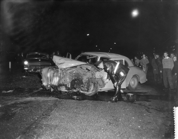 Schwarzes und weißes Foto eines auf der Straße gestrandeten Wagens mit mehreren Menschen in der Nähe und beleuchteter Hintergrund.