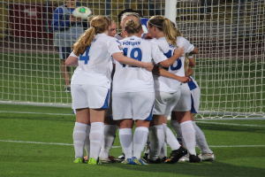 Gruppe von Frauenfussballspielerinnen in einheitlichen Uniformen auf dem Feld mit einem Netz und einer Person mit einem Ball im Hintergrund.