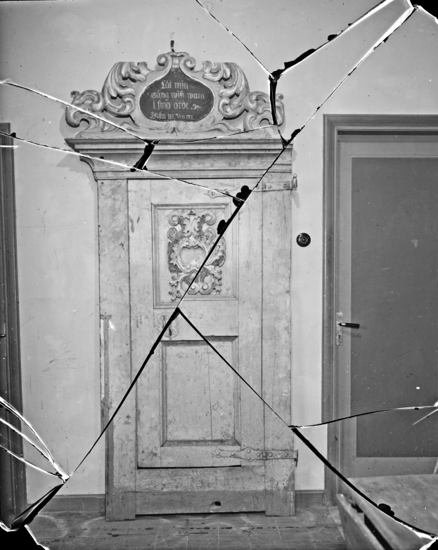 Ein Schwarz-Weiß-Foto einer kaputten Tür in einem Raum, mit zersplittertem Glas drumherum und einer Wand im Hintergrund.