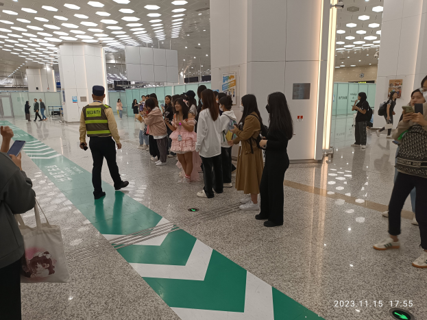 Menschen in einer Reihe an einem Flughafen-Sicherheitscheck-in, einige halten Taschen und Mobiltelefone, mit Säulen und Deckenlampen im Hintergrund.